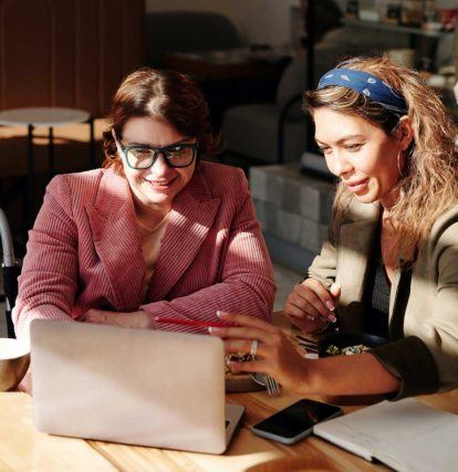 two women sitting at a table looking at a laptop