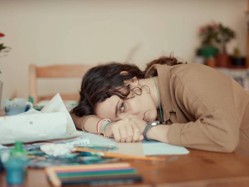 Person resting their head on a table with art supplies and papers, appearing tired or discouraged.