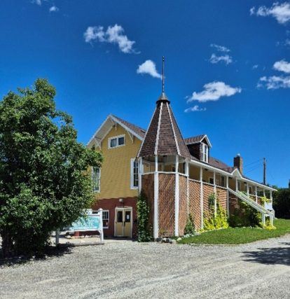 Large yellow historic building with a distinctive turret, wraparound porch, trees, and a bright blue sky.