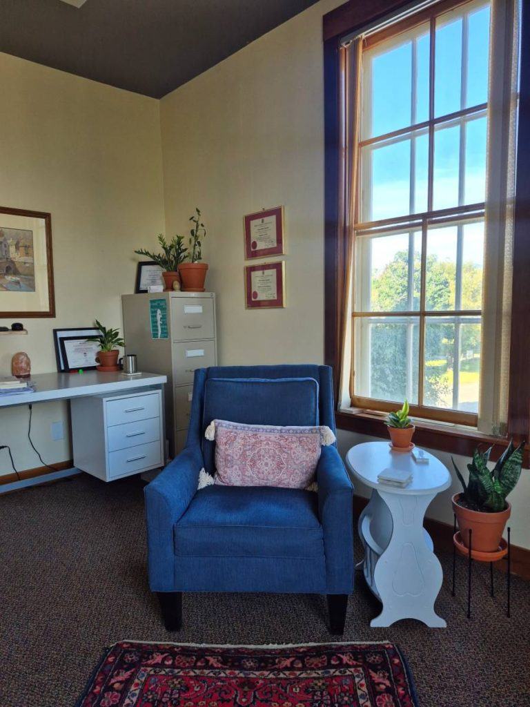Cozy therapy office with a blue armchair, side table, plants, desk, and large sunlit window.